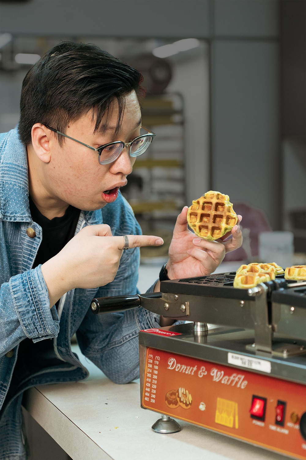 A person wearing glasses and a denim jacket points excitedly at a small Belgian waffle held in their hand next to a waffle maker labeled “Donut & Waffle,” possibly the best waffle machine for breakfast lovers.
