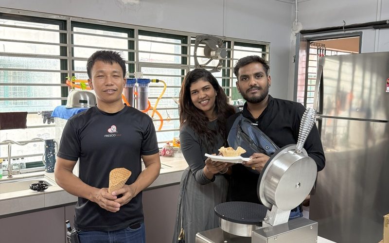 Three people stand in a kitchen beside a waffle cone maker and the best ice cream machine; one holds a cone, another has waffles on a plate, while the third stands between them, smiling at the camera.