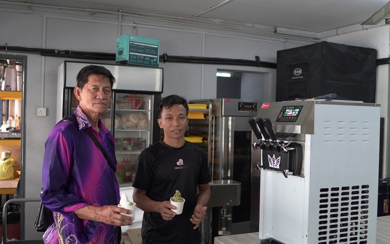 Two men stand in a room with an ice cream maker, holding cups of soft serve and looking at the camera, showcasing one of the popular types of ice cream machines.