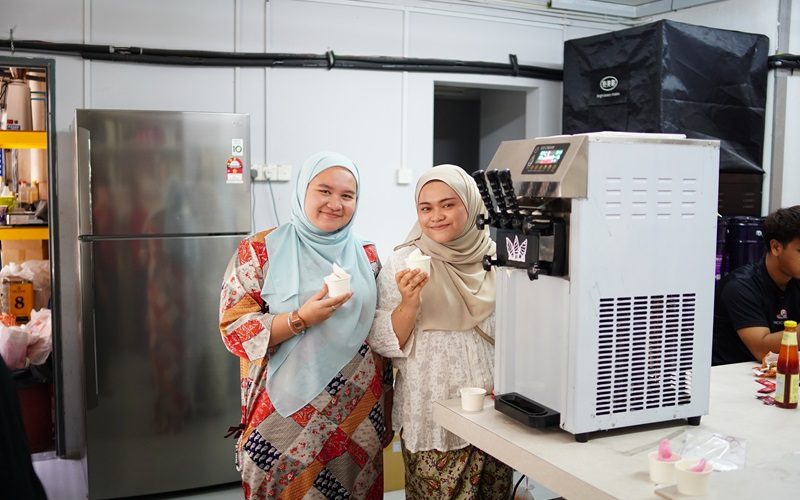 Two women in hijabs stand beside an ice cream maker, holding cups of soft-serve and smiling in a kitchen setting—showcasing popular types of ice cream machines used in Malaysia.