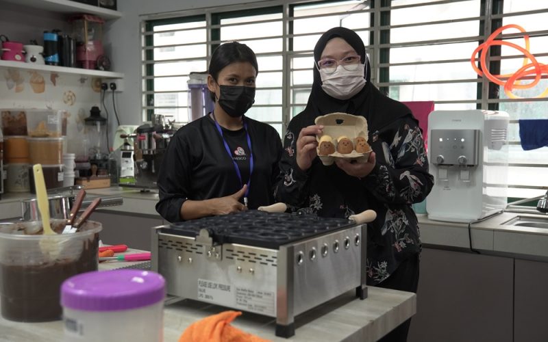 Two women wearing masks stand in a kitchen; one holds a carton with four egg-shaped pastries made with an Egg Waffle Machine. Cooking equipment and ingredients are visible on the counter, perfect for those looking to Start Small Food Business ventures.