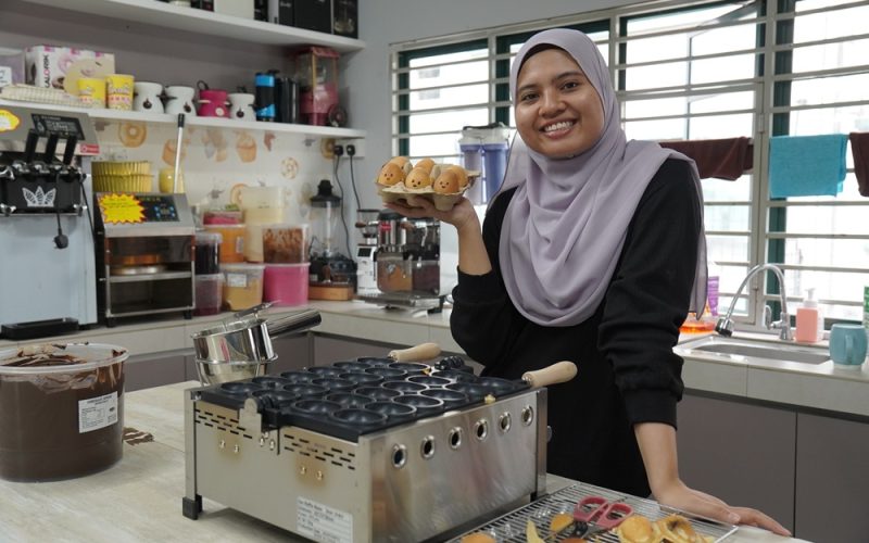 A woman in a hijab stands in a kitchen, smiling and holding cartoon-shaped baked goods made with an Egg Shape Waffle Maker, with baking equipment and supplies on the counter—perfect inspiration to Start Small Food Business dreams.