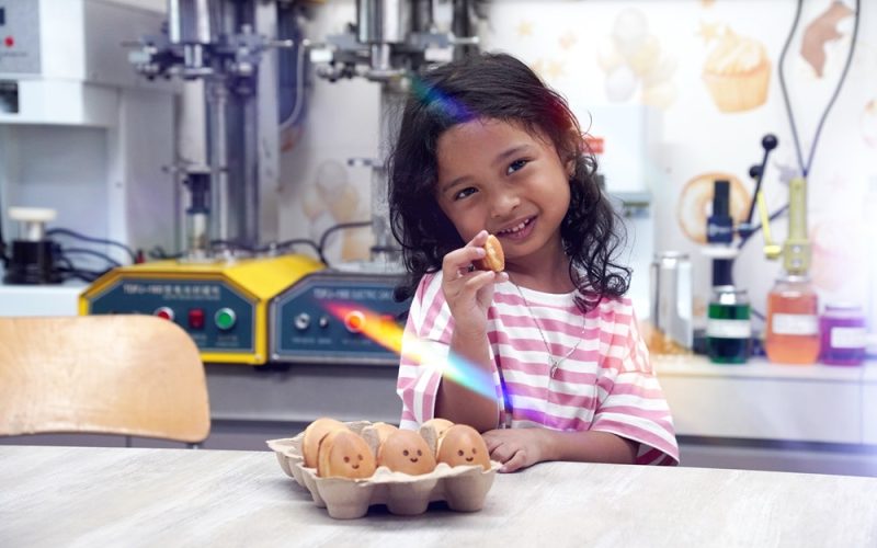 A young girl sits at a table holding a cookie, smiling with a tray of bear-shaped treats baked using the Fresco Malaysia Waffle Machine in front of her, in a cozy kitchen or bakery setting.