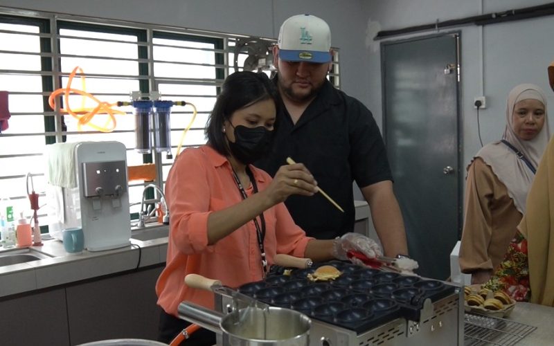 A woman uses chopsticks to cook food in a takoyaki pan, possibly exploring ideas for a profitable snack business, while others observe in a kitchen setting.