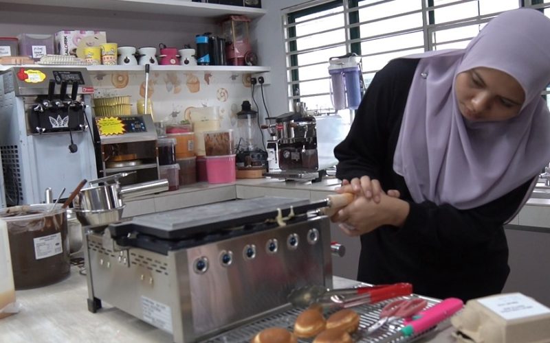 A woman in a hijab cooks at a countertop griddle in a kitchen, surrounded by baking tools, ingredients, and her Fresco Malaysia Waffle Machine—perfect for anyone looking to start a small food business or launch a profitable snack business.