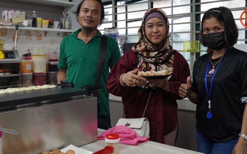 Three people standing in a kitchen, one holding a plate of food. Counter displays various items, including eggs. They are smiling, with one person wearing a mask.