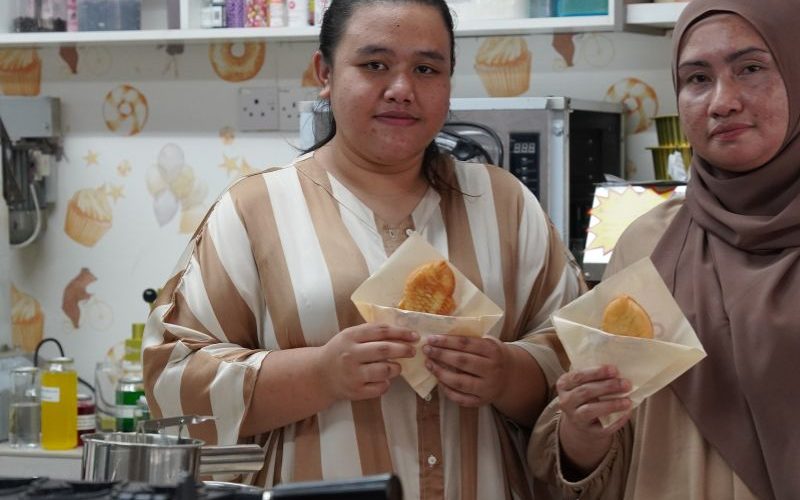 Two people in a kitchen holding pastries. Shelves with jars and bottles are visible in the background.