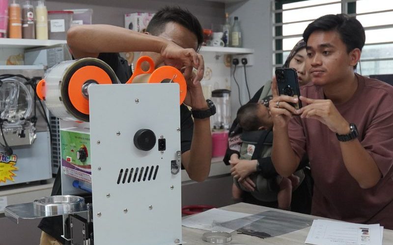 People observing a machine demonstration in a kitchen. One person records with a phone, while another adjusts the machine. Shelves with items are in the background.